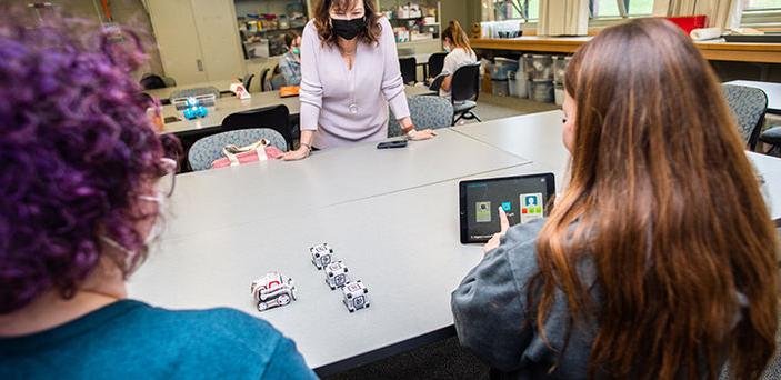 Professor Jeanne Ingle leans over a classroom table while 2 students sitting at the table work with an ipad and electronic blocks and vehicle